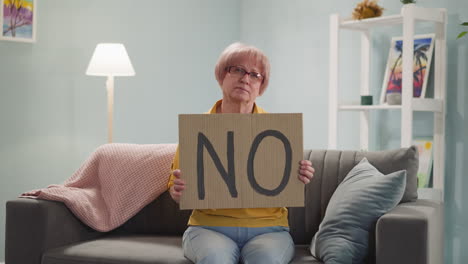 Upset-senior-woman-holds-placard-with-word-NO-on-sofa