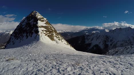 snowy-mountain-top-with-mountain-range-in-background-covered-in-clouds-under-a-blue-sky-with-shadow-of-the-cameraman-at-the-end-of-the-clip