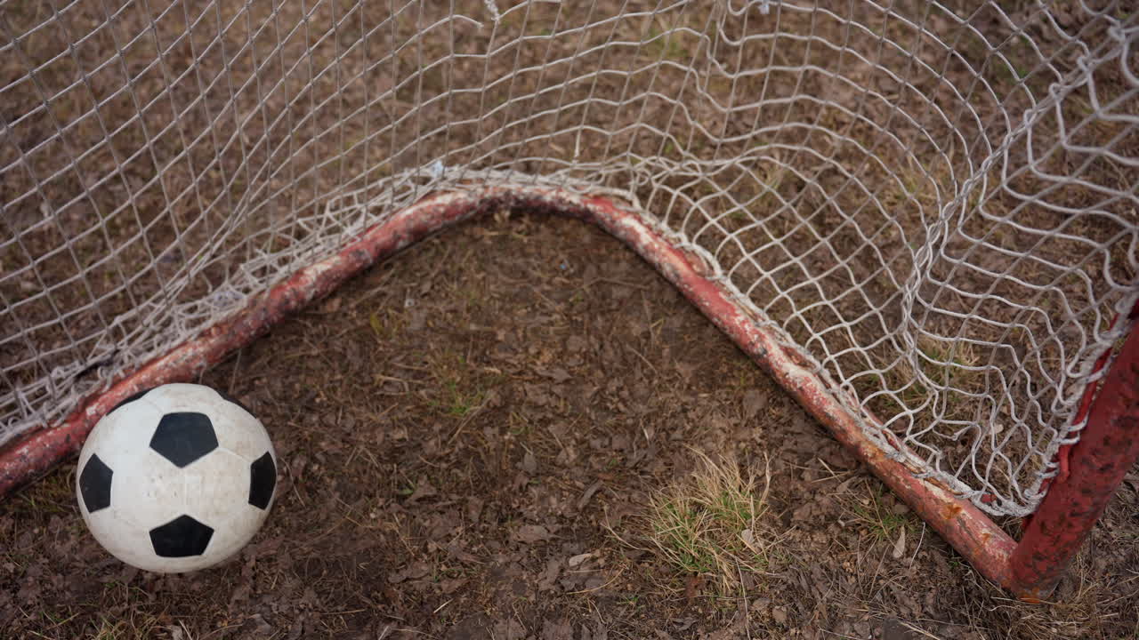 Вид сверху на мяч, попадающий в угол сетки, удар сотрясает сетку над утрамбованным грунтом, разлетаются частицы земли и вытоптанной травы, вид изнутри ворот с простой рамкой сетки, непосредственное ощущение гола