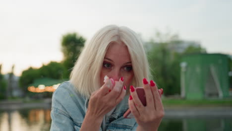 elegant woman sitting on riverside bridge in park during sunset uses compact mirror and white cloth to gently remove bright pink paint from nose after playful painting activity outdoors