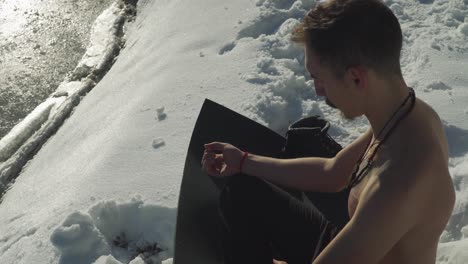 Man-sitting-on-black-yoga-mat-in-snow-with-no-shirt-breathing-heavily-to-prepare-mind-and-body-for-frigid-plunge-into-frozen-mountain-lake