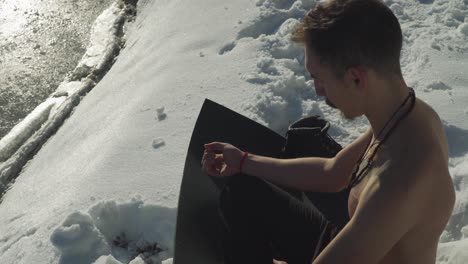 hombre sentado en una alfombra negra de yoga en la nieve sin camisa respirando pesadamente para preparar la mente y el cuerpo para sumergirse en un lago helado de montaña