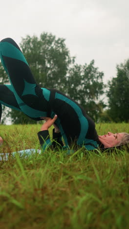 mujer de mediana edad acostada en una alfombra de yoga practicando la postura del puente con las manos apoyando su cintura en un vasto campo de hierba bajo cielos nublados, con árboles alineados en la distancia