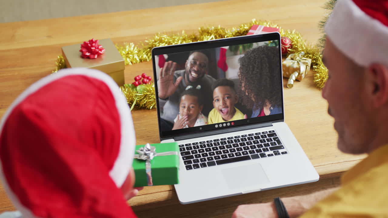 padre y hijo caucásicos con sombreros de santa usando una computadora portátil para una videollamada de navidad con la familia en la pantalla