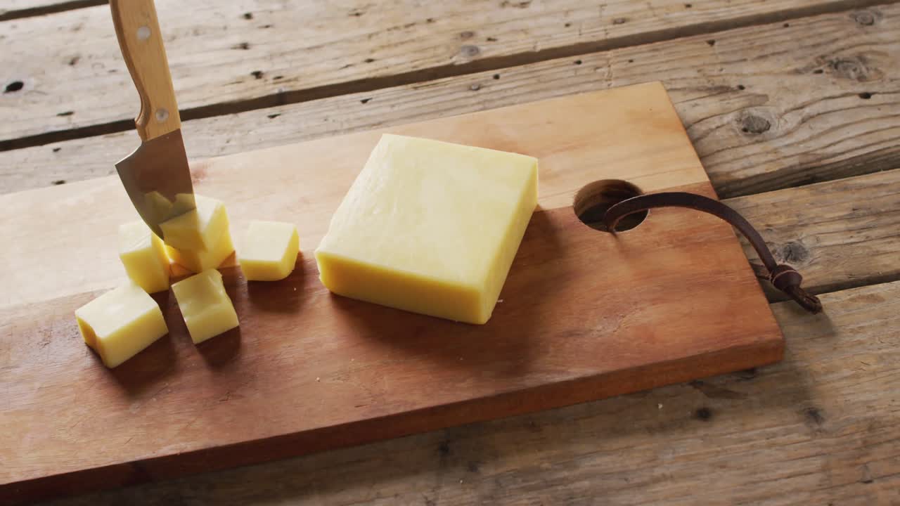 video de un bloque de queso y un cuchillo en cubos de queso en una tabla de madera y una mesa rústica