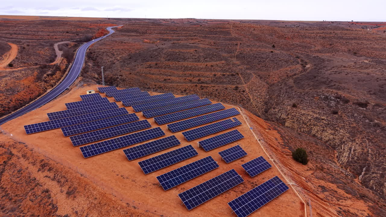 Luftfoto af nettilsluttet solcelleanlæg på et nivelleret plateau, med moduler i fast hældning, inverterstationer og perimeterhegn integreret med adgangsvejinfrastruktur.