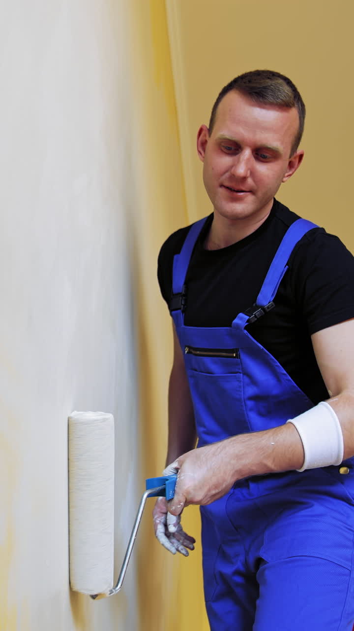 Portrait of a painter in action. Young man builder in blue overalls decorating room with a paint roller in the apartment. Room makeover. Vertical video