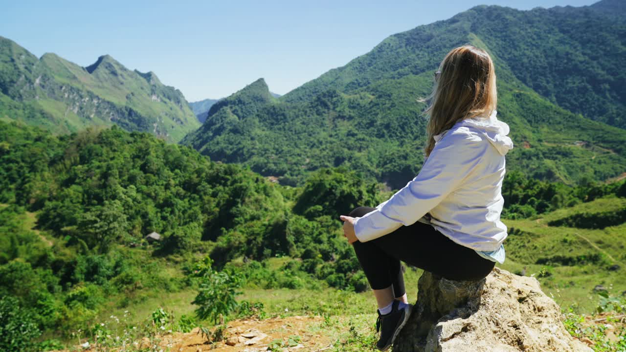 카우카스 여성 관광객이 산 꼭대기의 가장자리에 앉아 자연과 산의 풍경을 바라보고 있습니다.