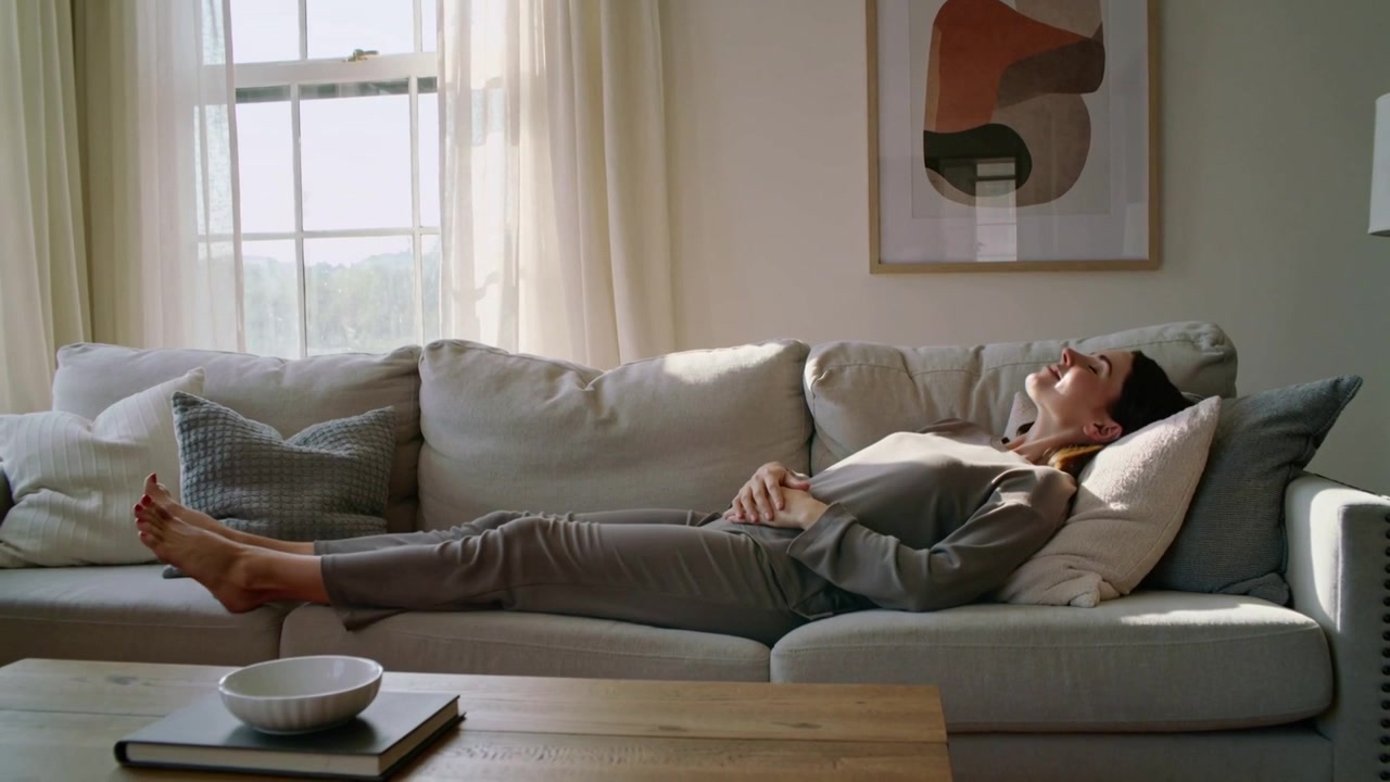 Sunlight drawing woman wearing soft grey loungewear reclining on sofa, seeking rest with window