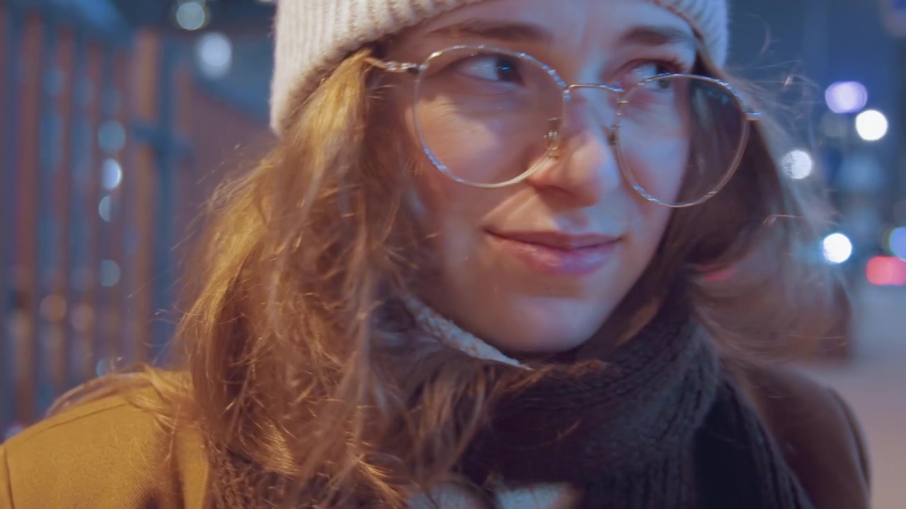 Nahaufnahme-Porträt einer Frau mit Mütze in der Nacht, mit Brille und Schal, umgeben von weichen Bokeh-Stadtlichtern. Sie trägt einen gemütlichen Wintermantel, zeigt ein subtiles Lächeln und einen nachdenklichen Ausdruck, in filmisch warmen Tönen.
