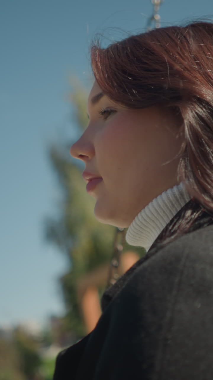 primer plano de una joven balanceándose al aire libre, el cabello balanceándose suavemente con la brisa, con una vegetación vibrante, edificios urbanos borrosos y un cielo azul brillante