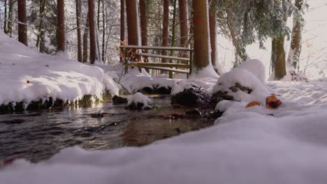 Ein-Bach-Im-Winter,-Der-Unter-Einer-Kleinen-Brücke-In-Einem-Wald-Fließt,-Mit-Sonnenstrahlen,-Die-Durch-Die-Zweige-Brechen