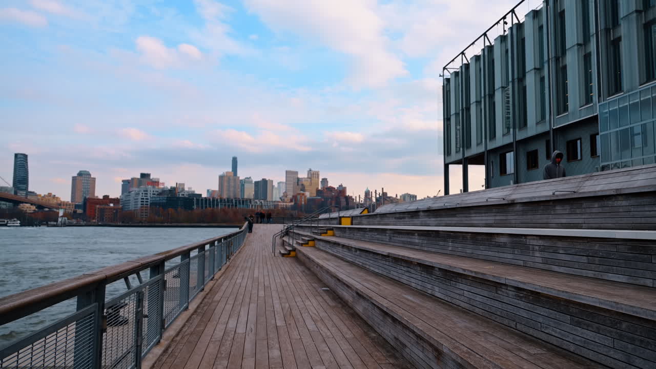 New York, USA, 5 dicembre 2025: Pontile in legno sul lungofiume e posti a sedere con skyline di Brooklyn. Ampia veduta di un moderno pontile in legno sul lungofiume e area con posti a sedere a gradoni con vista sul fiume e sul lontano skyline di Brooklyn al crepuscolo.