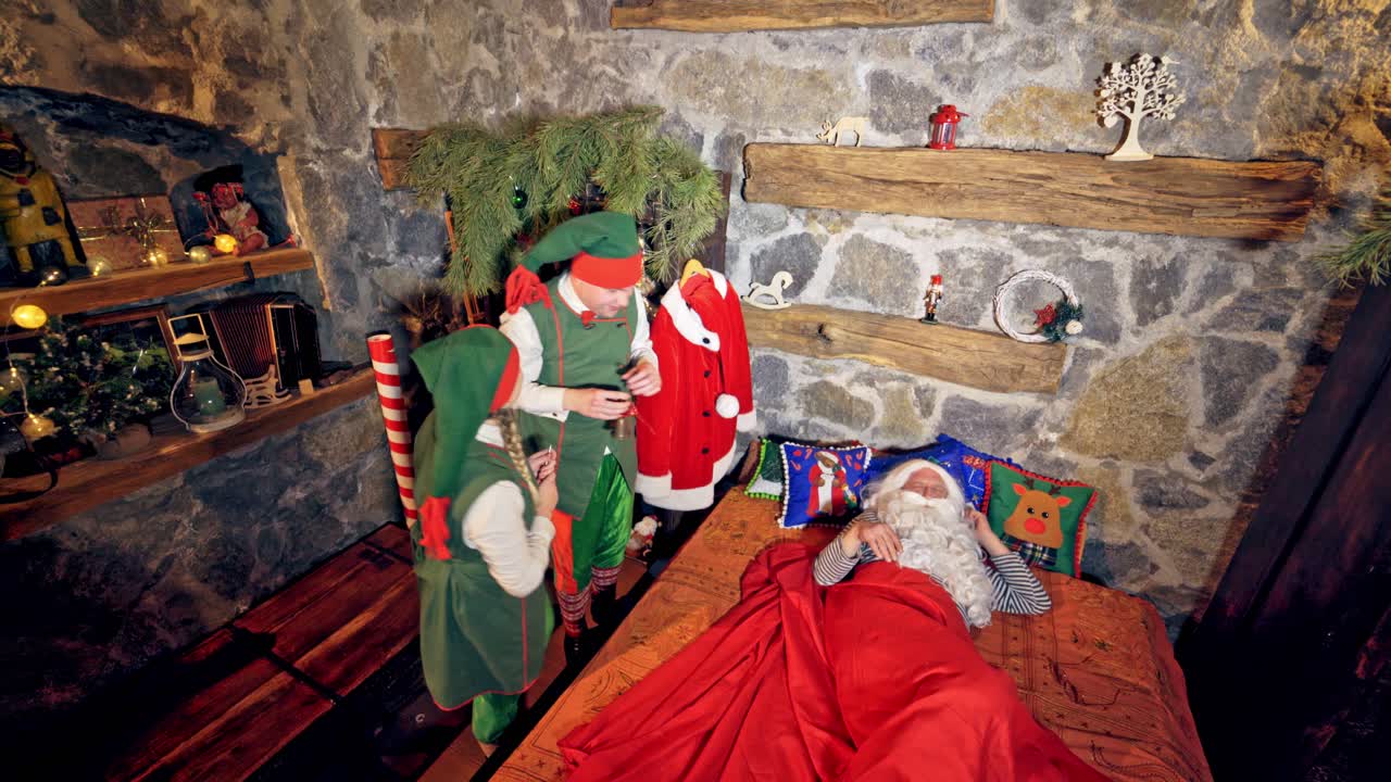 Santa Claus is sleeping in specially decorated room in his bed. Hand under cheek. Elves are ringing bell and touching him. Christmas spirit concept close up video. Video from above.