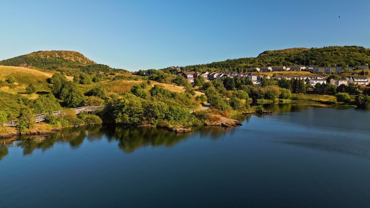 A drone glides along Quidi Vidi Lake's calm shoreline, capturing reflections of colourful houses, lush green trees, and serene water on a bright day
