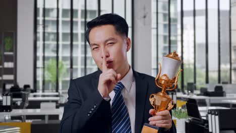 Close Up Of Asian Business Man In A Suit And Tie With A Gold Medal And Trophy Making Shh Gesture Telling It Is A Secret To Win As The First Winner In Office