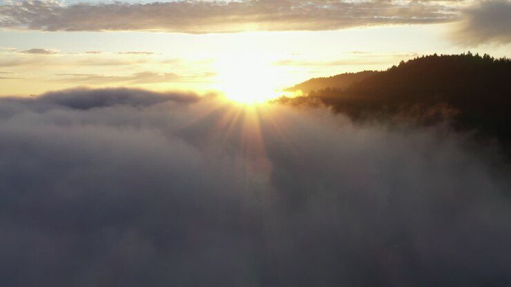 Imágenes aéreas de dron de rayos de sol asomándose sobre una niebla mágica y cimas de montañas al atardecer