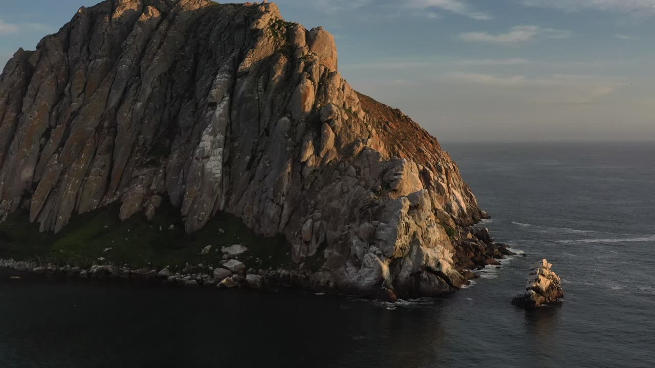 Panoramica aerea con drone attorno a una formazione rocciosa unica su un'isola durante il tramonto, mentre il sole si riflette sulla roccia a Morro Bay, California.