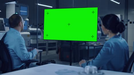 reunión de oficina: ingeniera confiada habla con el gerente de proyecto, viendo la televisión de pizarra digital interactiva que muestra la pantalla verde de la pantalla de la llave de croma. fábrica moderna con maquinaria