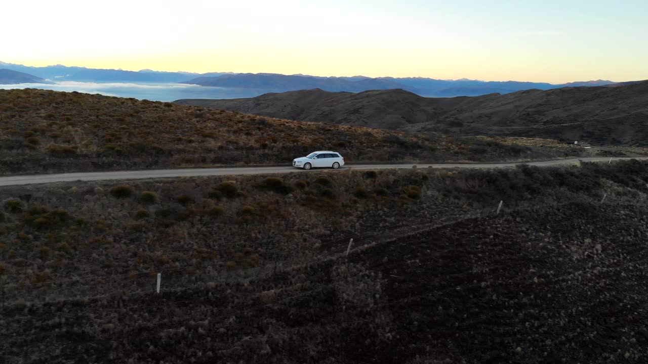 Carro branco percorre uma crista na Thomson Gorge Road em Central Otago ao pôr do sol, com vastas vistas de vales e montanhas ao longe