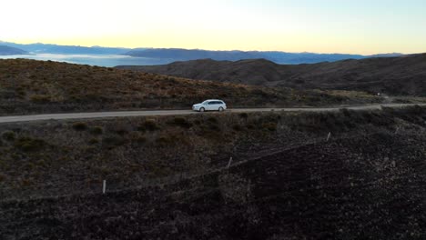 White-car-drives-along-a-ridgeline-on-the-Thomson-Gorge-Road-in-Central-Otago-at-sunset-with-vast-vistas-of-valleys-and-mountains-in-the-distance