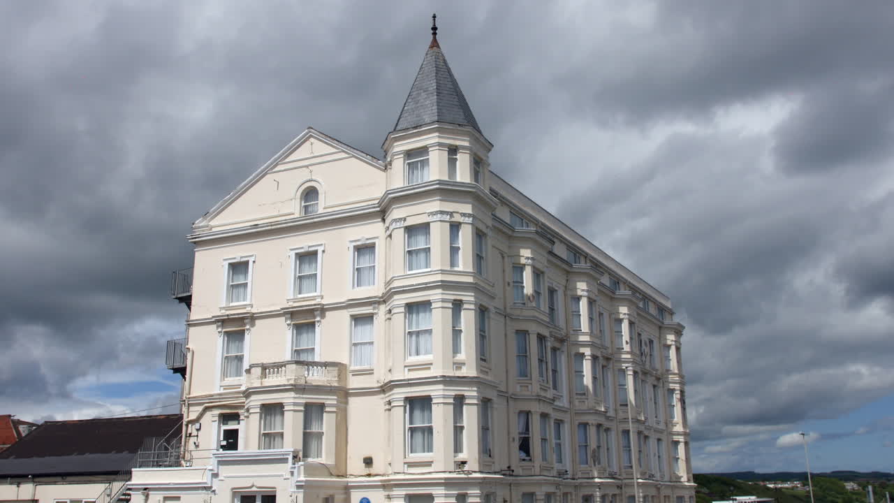 A building with Victorian architectural design stands under a cloudy sky, featuring tall windows, ornate details, and a conical turret. Dark storm clouds are gathering in the background
