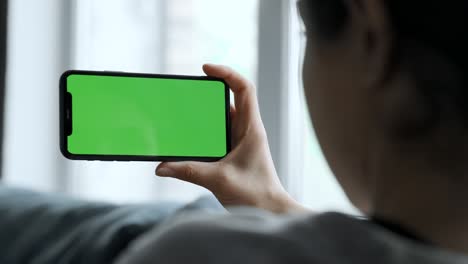 여성의 손은 녹색 화면의 스마트폰을 들고 소셜 네트워크, 영화, 비디오를 보고, 편안한 방의 소파에 누워 있습니다.