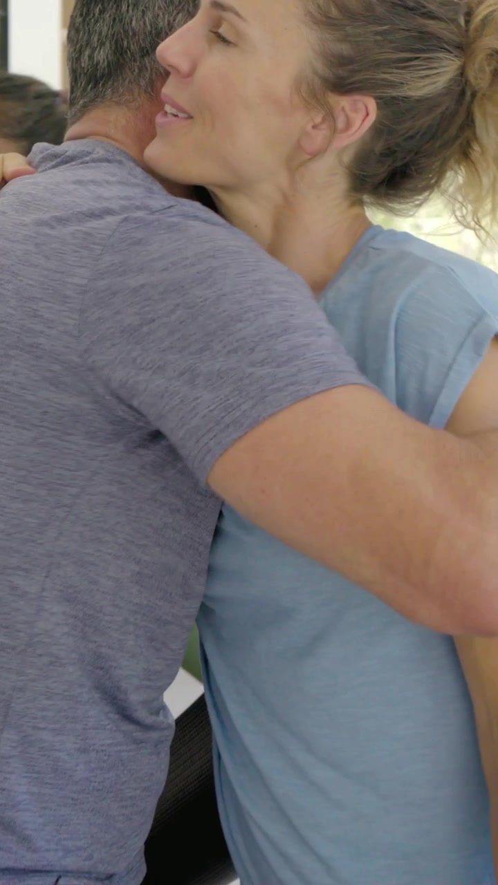 Video verticale: Donna con maglietta blu che entra in salotto portando un tappetino, salutando gli amici per lo yoga.