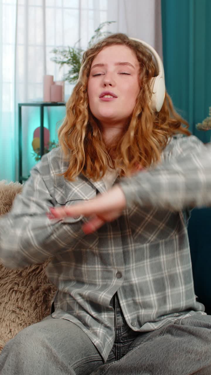 Woman sitting on sofa wearing headphones dancing with closed eyes enjoying favorite relaxing music