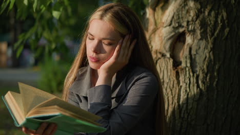 lady in grey clothing seated under tree, head resting on left hand while reading book, looking moody, sunlight softly illuminates her face, with background slightly blurred, featuring greenery