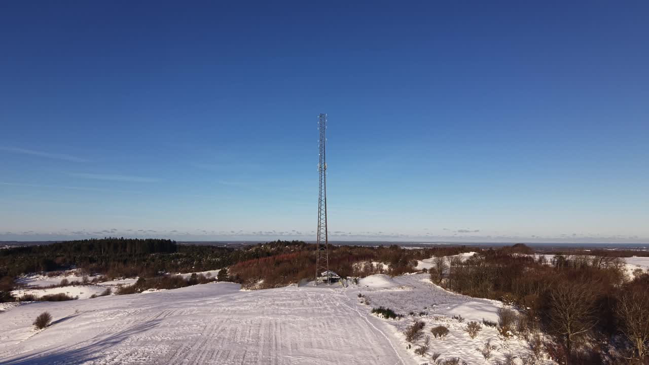 A sweeping drone shot captures a tall lattice communication tower standing atop a snow-covered hill. The scene features a clear blue sky, distant forests, and a peaceful wintry horizon