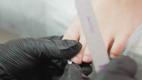 close-up view of nail technician in black gloves carefully trimming client's toenail while holding nail file during pedicure procedure, emphasizing precision, hygiene, and professional care