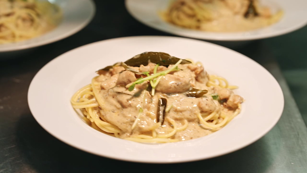 Inquadratura macro statica della mano guantata di uno chef che grattugia finemente parmigiano su spaghetti al pollo cremosi appena impiattati, guarniti con micro-ortaggi ed erbe aromatiche