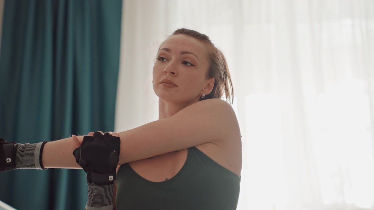 Donna che allunga il braccio nel soggiorno di casa, tatuaggio visibile, guanti senza dita e fascia per il polso, respiri calmi e misurati e raggio di movimento controllato, morbida retroilluminazione attraverso le tende, routine di pre-allenamento focalizzata