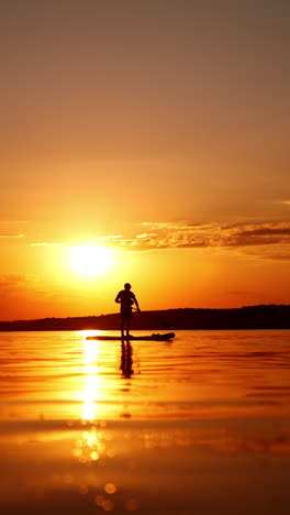 black silhouette of a male standing on the paddle board at sunset. man uses an oar to move by the river. low angle view. vertical video.