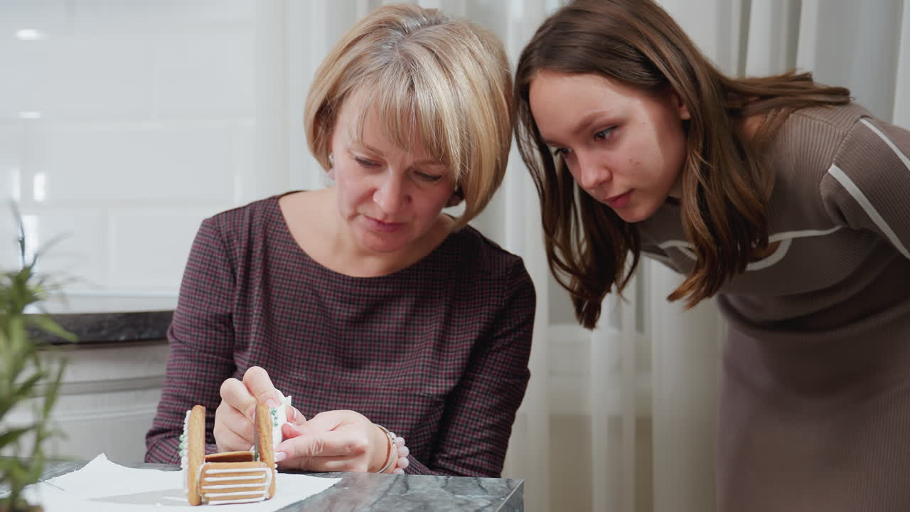 딸은 조심스럽게 그녀의 어머니를 지켜보고 그녀가 조심스레 진저브레드 하우스 케이크에 아이싱을 적용합니다. 전면에 부분적으로 보이는 녹색 식물과 배경에 부 요소가 있습니다.