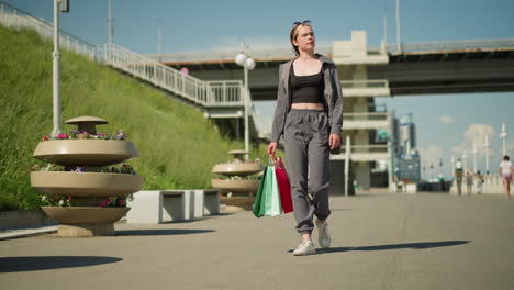 lady walks holding shopping bags in her right hand, wearing open grey clothing, people walk in the background near a flower vest, with a visible bridge and cars driving overhead