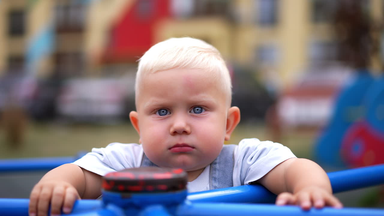 Serious cute blond baby boy stands on the carousel. Grey-haired Caucasian toddler at the playground. Close up. Blurred backdrop.