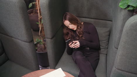 upward angle shows young woman dressed in black seated in cushioned booth, head tilted and eyes focused on smartphone. background features brick wall and potted plant in black vase for cozy ambiance