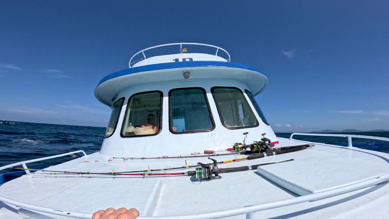 Handheld camera captures fishing rods on boat deck under bright daylight, gentle waves, clear sky