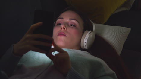 Mujer-Con-Auriculares-Inalámbricos-Tumbada-En-El-Sofá-De-Casa-Por-La-Noche-Transmitiendo-Música-O-Viendo-Una-Película-En-Un-Teléfono-Móvil-4