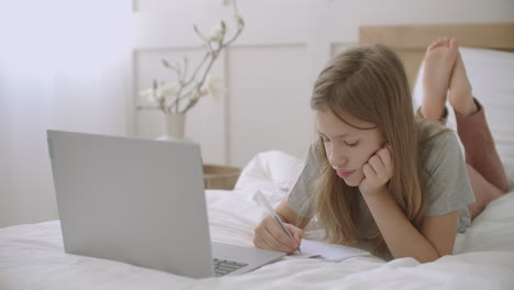 bored schoolgirl is drawing in copybook during online lecturing of teacher on laptop homeschooling and e-learning of children