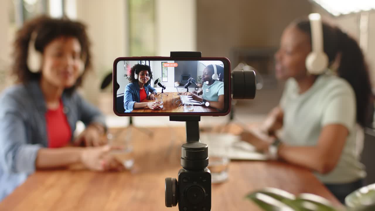 Diverse co-hosts tapping smartphone, using microphones and talking while recording podcast on table