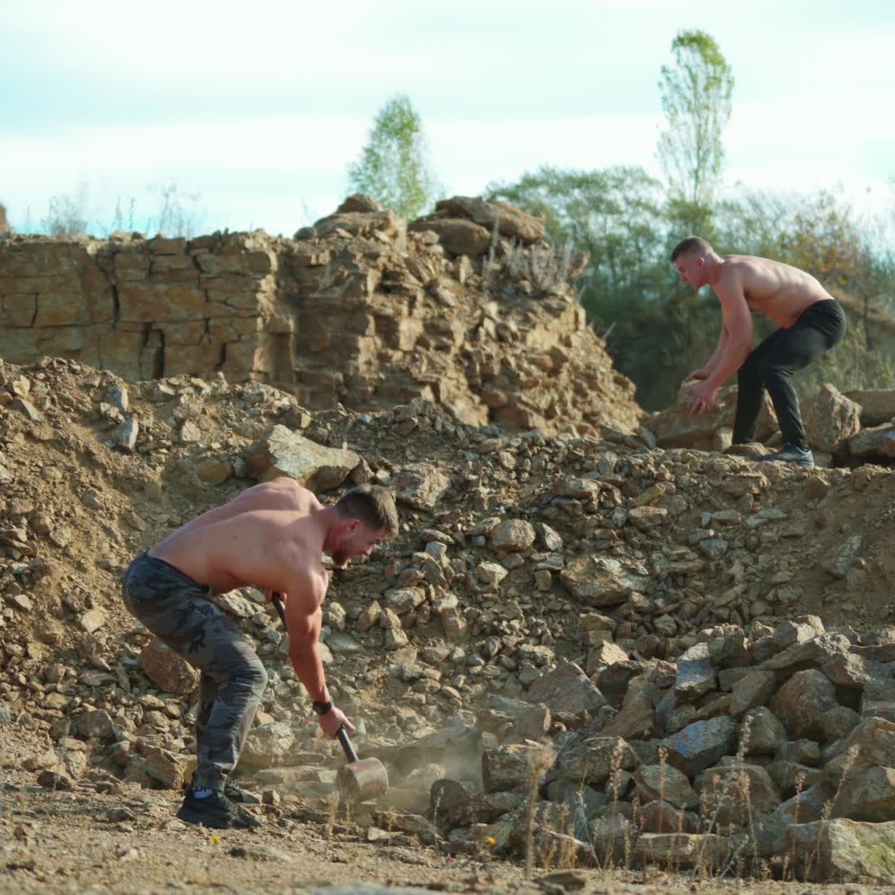 Two sportsmen breaking stones outdoors. Shirtless guy with a hammer on the rocky background. Athlete men training their body with hard exercises.