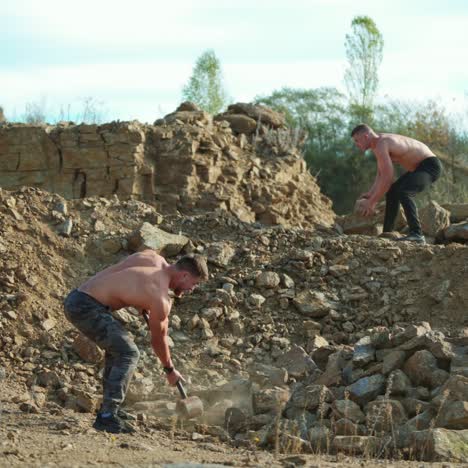 two sportsmen breaking stones outdoors. shirtless guy with a hammer on the rocky background. athlete men training their body with hard exercises.