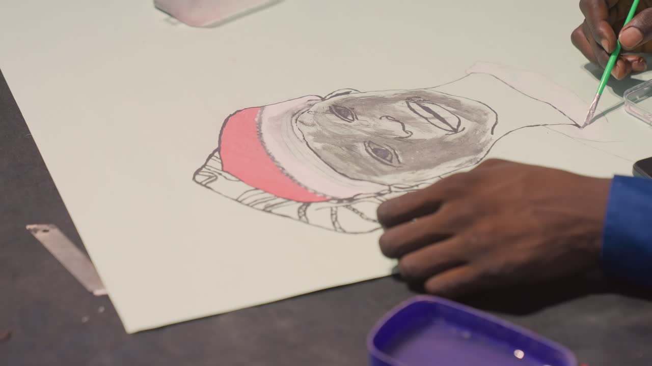 Pintura de retrato vista de cima com mão de pele escura, aplicando camada rosa a um gorro de malha, pincel de cerdas verdes movendo-se em pinceladas medidas, mesa de estúdio com bandeja de pigmentos e smartphone, atmosfera de oficina urbana.