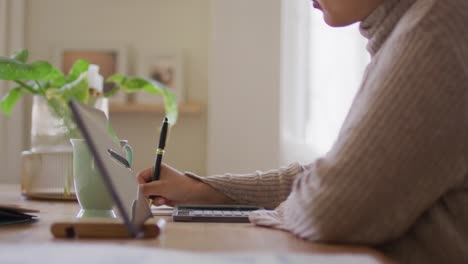 video de una mujer biracial con hijab sentada en un escritorio en casa trabajando en una tableta