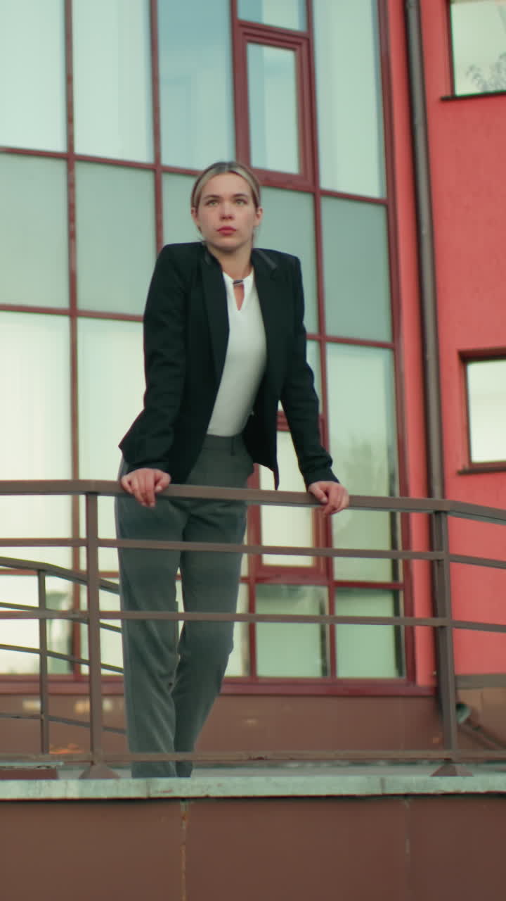 Young lady in black blazer resting hand on iron railing gazes thoughtfully into distance beside lamp post, with modern glass building and red wall background