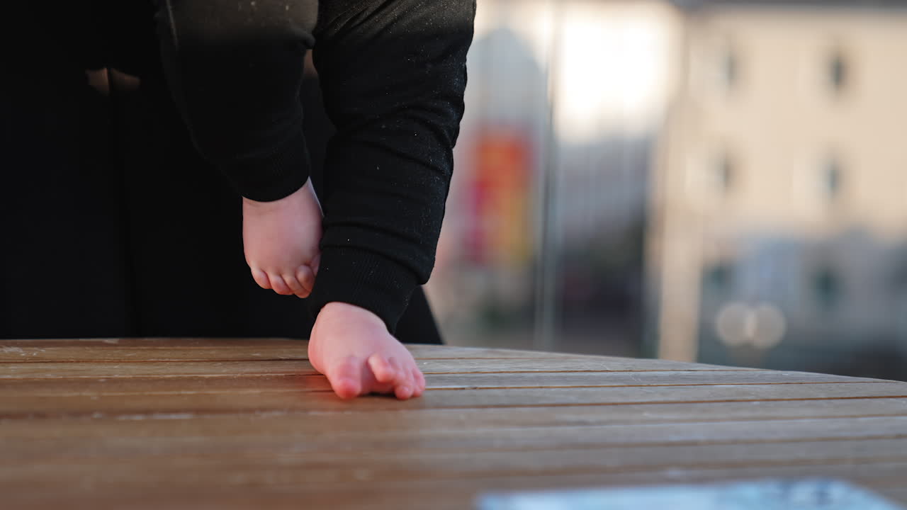 Tiny bare feet of a baby on the wooden surface. Kid wearing black pants stepping by the table. Blurred backdrop.