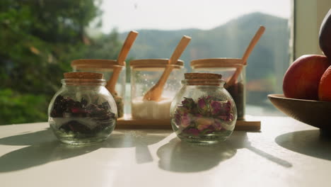 tabletop displays components for herbal tea. glass containers filled with dried flowers and herbs against window with view of nature. natural drink